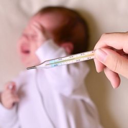 Eine Frau hält ein Fieberthermometer, im Hintergrund ist ein Baby zu sehen.